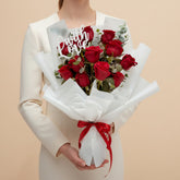 A bouquet of red roses wrapped in white tissue paper with a red ribbon and a white script 'Love' sign, displayed on a white pedestal against a warm beige background.