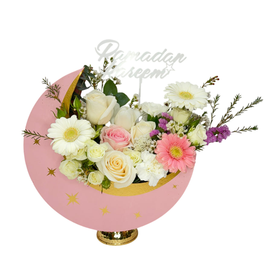 A crescent moon-shaped floral arrangement with pastel roses, gerberas, and mixed flowers in a pink and gold base, adorned with a "Ramadan Kareem" stick
