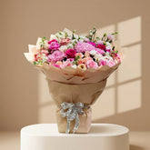 A large floral bouquet with pink, magenta, peach, and white flowers wrapped in tan paper and tied with a white ribbon, displayed on a white pedestal against a white wall.