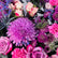 A vibrant floral arrangement in a white cylindrical container from Viva Flora, featuring prominent purple and pink flowers including chrysanthemums, roses, carnations, and blue thistles on a light background.