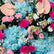 A vibrant floral arrangement featuring light blue hydrangeas, bright pink carnations, peach anthuriums, and small white baby's breath on a white background.