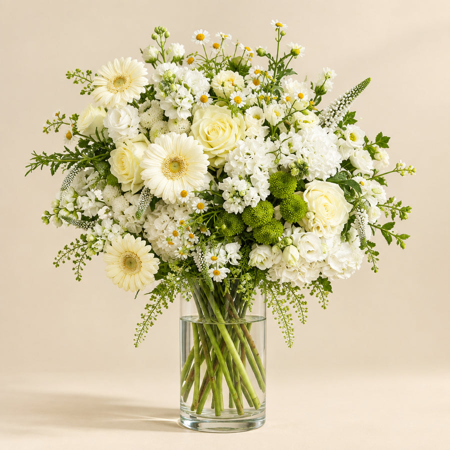 A vibrant floral bouquet featuring white daisies, roses, carnations, and green foliage arranged in a clear glass vase against a solid beige background.