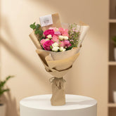 A wrapped bouquet of pink, red, and white roses with green leaves, wrapped in brown kraft paper and tied with twine, displayed on a white cylindrical pedestal in a softly lit indoor setting.