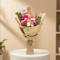 A wrapped bouquet of pink, red, and white roses with green leaves, wrapped in brown kraft paper and tied with twine, displayed on a white cylindrical pedestal in a softly lit indoor setting.