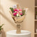 A wrapped bouquet of pink, red, and white roses with green leaves, wrapped in brown kraft paper and tied with twine, displayed on a white cylindrical pedestal in a softly lit indoor setting.