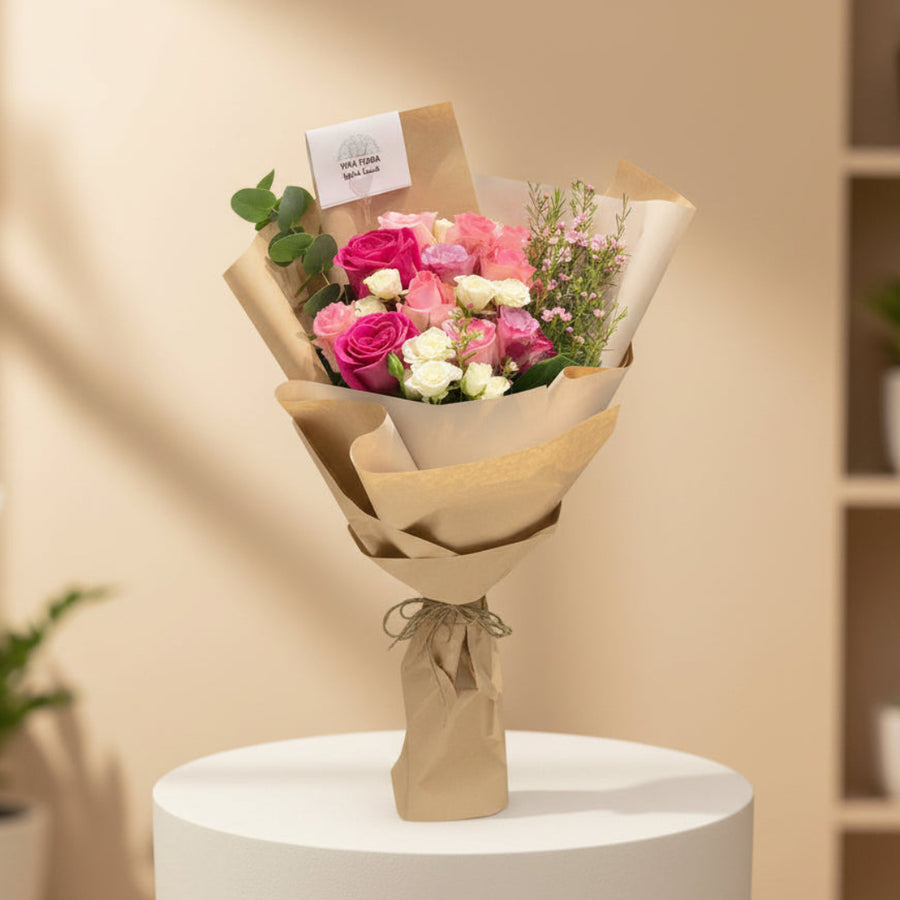 A wrapped bouquet of pink, red, and white roses with green leaves, wrapped in brown kraft paper and tied with twine, displayed on a white cylindrical pedestal in a softly lit indoor setting.