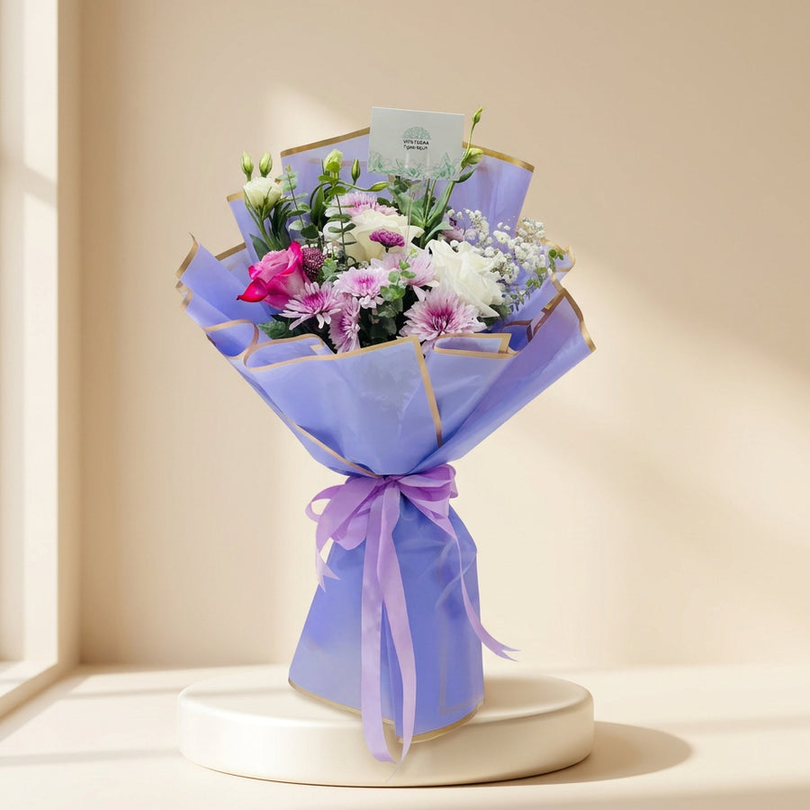 A vibrant flower bouquet wrapped in light purple paper with gold trim and tied with a matching purple ribbon, held by a person's arm in a brightly lit indoor setting.