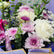 A vibrant flower bouquet wrapped in light purple paper with gold trim, featuring white and pink roses, purple chrysanthemums, and green foliage. A card for 'VIVA FLORA' is visible at the top.