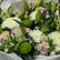 A large floral bouquet wrapped in white tissue paper, featuring white and light pink flowers with green stems and buds, placed on a light wood floor in front of a dark glass door.