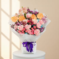 A vibrant floral bouquet featuring pink roses, orange anthuriums, purple hydrangeas, and pink carnations, wrapped in white tissue paper with a purple ribbon bow, displayed on a white marble surface.