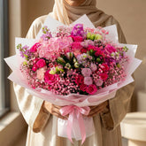 A vibrant pink and red floral bouquet wrapped in white and pink tissue paper, tied with a pink ribbon, displayed on a white pedestal against a warm beige background.