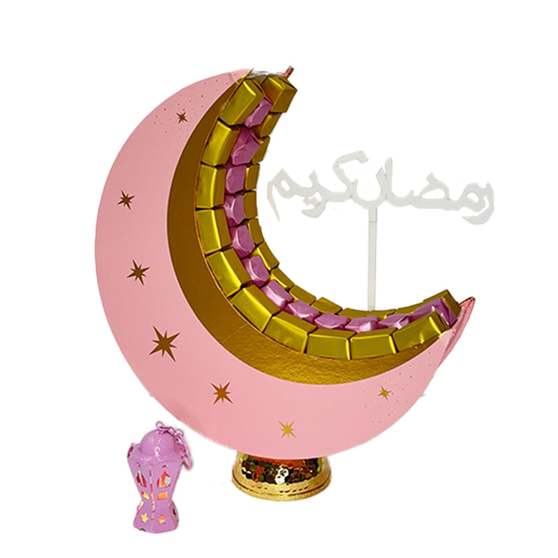  A crescent moon-shaped chocolate arrangement in pink and gold, decorated with stars, placed on a stand with a small lantern beside it.