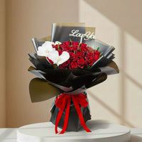 A bouquet arrangement featuring red roses, a white orchid, and a gold card with 'Love You' script, wrapped in black paper with a red ribbon, displayed on a round pedestal.