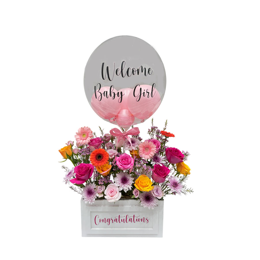 A white wooden box filled with pink flowers and a clear bubble balloon with the message "Welcome Baby Girl."