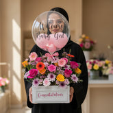 A floral congratulations arrangement in a white rectangular box with a large clear balloon reading 'Welcome Baby Girl' and pink accents, displayed on a white pedestal against a warm beige wall.