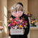 A floral congratulations arrangement in a white rectangular box with a large clear balloon reading 'Welcome Baby Girl' and pink accents, displayed on a white pedestal against a warm beige wall.