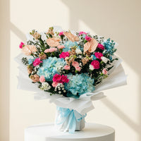 A vibrant bouquet of Natural flowers wrapped in white tissue paper and tied with a light blue ribbon, placed on a gray tiled floor in front of a blurred shop background.