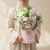 A floral arrangement in a round pink box with a pearl handle, displayed on a white round pedestal. The bouquet includes white and pale pink roses, green foliage, and small white filler flowers.