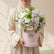 A floral arrangement in a round pink box with a pearl handle, displayed on a white round pedestal. The bouquet includes white and pale pink roses, green foliage, and small white filler flowers.