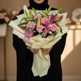A floral bouquet featuring pink roses, purple lilies, and green lily buds wrapped in cream paper and tied with a matching ribbon, displayed on a white pedestal against a beige background.