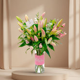 A bouquet of pink and green lilies in a clear glass vase with a pink label reading 'Maman', displayed on a white pedestal against a warm beige wall with soft window shadows.