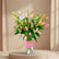 A bouquet of pink and green lilies in a clear glass vase with a pink label reading 'Maman', displayed on a white pedestal against a warm beige wall with soft window shadows.
