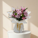 A beautiful bouquet of pink lilies, roses, and chrysanthemums wrapped in white paper with a gold ribbon.
