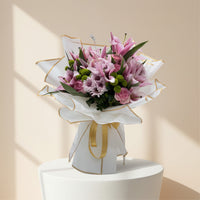 A beautiful bouquet of pink lilies, roses, and chrysanthemums wrapped in white paper with a gold ribbon.
