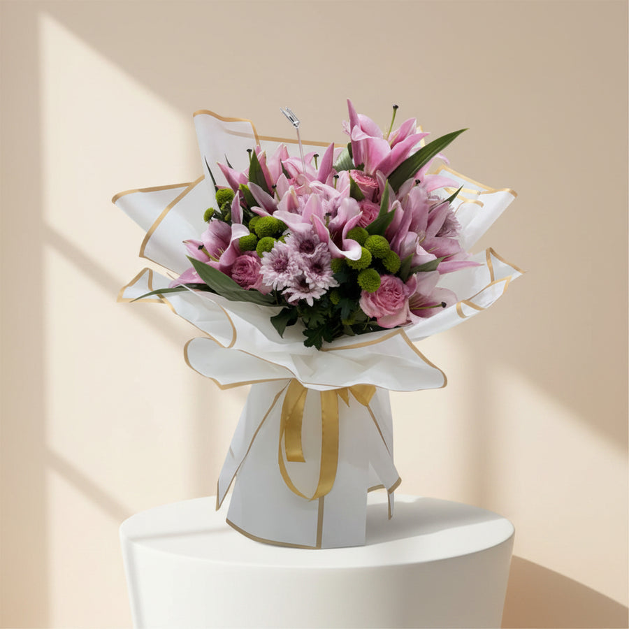 A beautiful bouquet of pink lilies, roses, and chrysanthemums wrapped in white paper with a gold ribbon.
