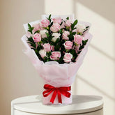 A bouquet of pink and white roses wrapped in light pink tissue paper with a red ribbon tied in a bow at the base, set against a light marble-patterned background.