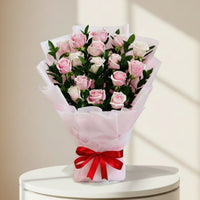A bouquet of pink and white roses wrapped in light pink tissue paper with a red ribbon tied in a bow at the base, set against a light marble-patterned background.