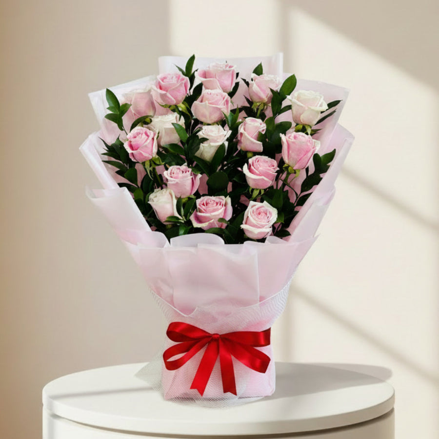 A bouquet of pink and white roses wrapped in light pink tissue paper with a red ribbon tied in a bow at the base, set against a light marble-patterned background.