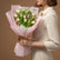 A bouquet of tulips wrapped in light pink paper and tied with a rustic twine bow, displayed on a white pedestal against a warm beige wall with soft window-shadow lighting.