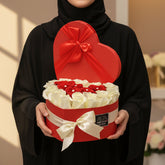 A red heart-shaped gift box with its lid propped open, filled with white rose petals and topped with a red heart, displayed on a white cylindrical pedestal against a light gray background.