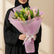A bouquet of pink lilies wrapped in pink tissue paper and a white ribbon, displayed on a round white table against a warm beige background with soft window-light shadows.