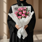 A bouquet of pink roses wrapped in white tissue paper with a white ribbon and printed card that reads 'best MOM ever,' displayed on a white pedestal against a warm beige background.