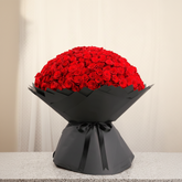 A large bouquet of red roses wrapped in black paper with a black ribbon printed with the brand name 'Rose World', displayed on a light gray speckled countertop against a white curtain background.