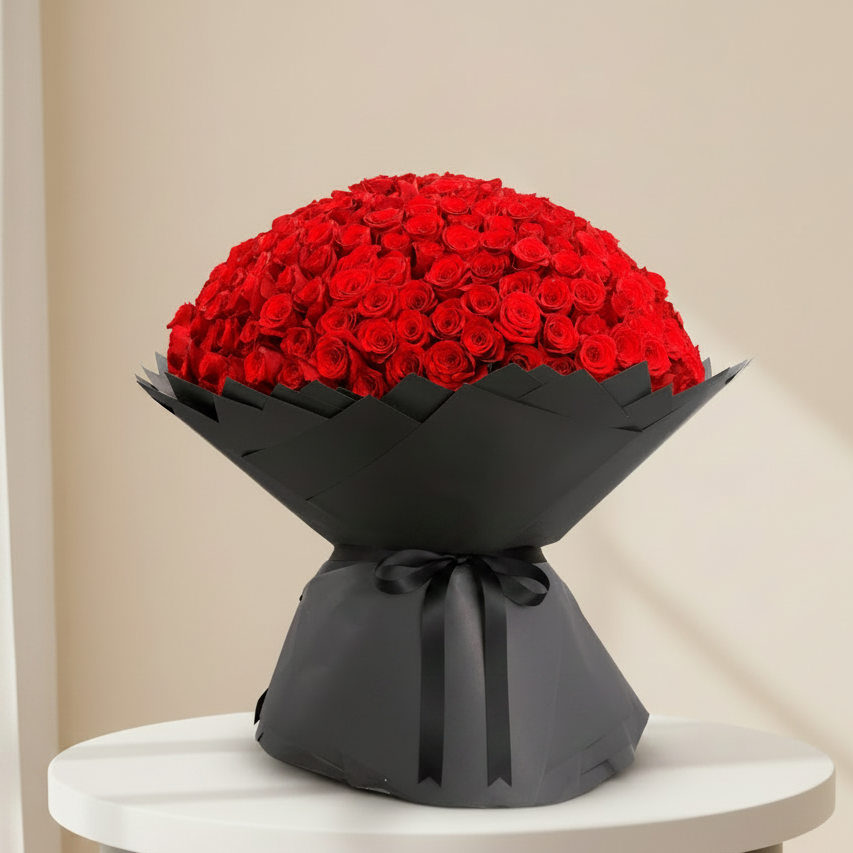 A large bouquet of red roses wrapped in black paper with a black ribbon printed with the brand name 'Rose World', displayed on a light gray speckled countertop against a white curtain background.