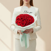 A bouquet of 50 red roses wrapped in pale mint green paper with a heart-shaped card that reads 'I Love you', displayed on a white round pedestal against a warm beige background with soft window-shadow lighting.