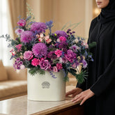 A vibrant floral arrangement in a white cylindrical box, featuring prominent purple and pink flowers with green foliage, displayed on a white pedestal against a white studio backdrop.