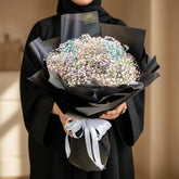 A bouquet of dried baby's breath flowers wrapped in black paper and tied with a white ribbon, displayed on a white pedestal against a warm beige wall with soft window shadows.