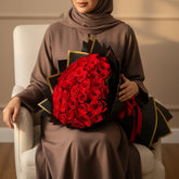 A large bouquet of red roses wrapped in black paper with gold edges, displayed on a white surface against a white background, with a second wrapped bouquet standing behind it.