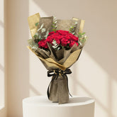A person in a black abaya and hijab holds a large bouquet of red roses wrapped in textured gold and brown paper with green eucalyptus leaves and a small card, photographed in a softly lit indoor setting.