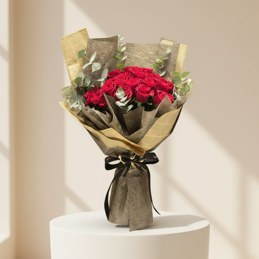 A person in a black abaya and hijab holds a large bouquet of red roses wrapped in textured gold and brown paper with green eucalyptus leaves and a small card, photographed in a softly lit indoor setting.