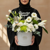 A floral arrangement in a round silver gift box with a gold logo, placed on a white pedestal against a warm beige background. The bouquet includes white lilies, white roses, white daisies, green chrysanthemums, green foliage, and a small lantern candle holder.