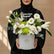 A floral arrangement in a round silver gift box with a gold logo, placed on a white pedestal against a warm beige background. The bouquet includes white lilies, white roses, white daisies, green chrysanthemums, green foliage, and a small lantern candle holder.