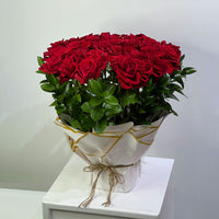 A large bouquet of 25 red roses with green leaves, wrapped in white paper with gold accents and tied with twine, displayed on a white pedestal against a light gray background.
