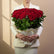 A large bouquet of red roses with green leaves, wrapped in white paper with gold ribbon accents, displayed on a white pedestal against a beige background with soft window shadows.