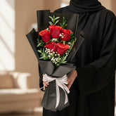 A bouquet of six red roses wrapped in dark green paper with a white patterned ribbon, displayed on a round white pedestal against a warm beige background.