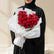 A heart-shaped bouquet of red roses wrapped in white tissue paper and tied with a white ribbon, displayed on a round white pedestal against a warm beige background with soft window shadows.